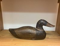 Side view of vintage wooden duck decoy showing paint wear and chipping on head and body, on a wood shelf.