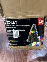 Original retail box of the NOMA 6.5 ft Mariposa Pre-Lit Colour-Changing Tree showing brand, size, and product details.