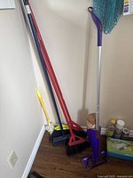 Photo showing four brooms and a purple Swiffer mop leaning against wall