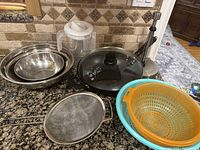 Photo showing three stainless steel mixing bowls nested, large black wok with glass lid, three strainers (orange, blue plastic, and stainless steel mesh), clear plastic drink container, and a standing paper towel holder on kitchen counter.