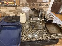 Top view of the assorted kitchen items including two French presses, two blue baking sheets, metal pot with glass lid, clear plastic drink container, assorted lids, and kitchen twine on granite countertop.