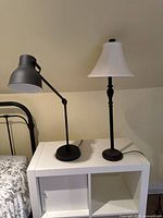 Two lamps on a white nightstand against a beige wall under a sloped ceiling. One lamp is an adjustable black metal desk lamp and the other is a taller lamp with a fabric shade.