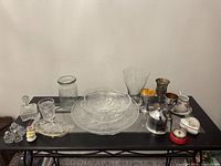 Overview of assorted decorative objects including glass bowls, vases, silver-plated cups, ceramic figurines, and small containers arranged on a table.
