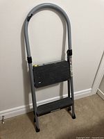 Front view of Cosco metal step ladder leaning against wall, showing two black steps and gray metal frame with arched handle.
