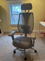 Front view of gray ergonomic office chair with headrest and mesh backrest in room with window and yellow walls.