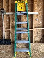 Front view of the Featherlite 6ft green and yellow step ladder leaning against a plywood wall with a snow shovel nearby.
