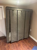Grey fabric portable wardrobe with zippered front cover positioned against wall on hardwood floor.