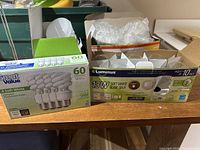 Two boxes of light bulbs on a table; left box Great Value 60W soft white, right box Luminus 13W soft white CFL bulbs with some bubble wrap and loose bulbs