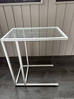 Side profile view showing white metal frame and glass top surface of the table on dark floor against light wall.