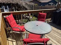 Photo of round glass outdoor patio table with three red cushioned metal chairs on wooden deck