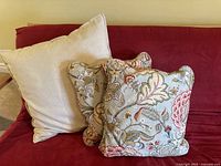 Three pillows arranged on a red couch, including one plain cream-colored pillow and two decorative floral-patterned pillows.