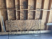 Black wrought iron queen-sized headboard displaying elegant floral and spiral patterns, photographed against a wooden wall background.