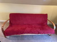 Front view of red cushion convertible futon sofa with light wood frame against beige wall and carpet flooring.
