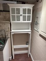 Front view of the white over the toilet storage unit showing the two glass-paneled cabinet doors and lower open shelf section.