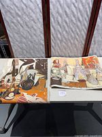 Two abstract oil paintings on canvas rolled, placed side by side on table, showing full canvases with warm colors and still life motifs.