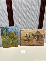 Two art pieces displayed side by side: African cloth art on wood and still life painting on board.