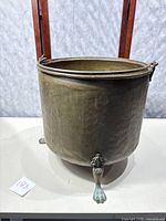 Front angled view of large round footed brass bucket with metal handle, showing hammered surface and claw foot legs.