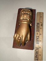 Close-up photo of the brass woman's hand door knocker mounted on a wooden plaque with a ruler for size reference.