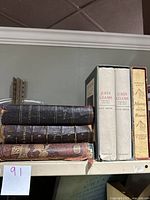 Six antique books arranged on a shelf including three leather-bound St. Nicholas volumes, two John Adams volumes in white slipcase, and one Mutiny on the Bounty book with yellow spine.