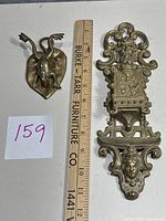 Two brass items including a wall hanging match holder and a frog clip placed side by side with measurement ruler.