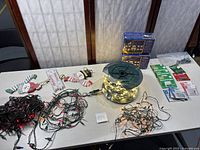 Overview of the lot on a table showing all components: boxed icicle lights, outdoor spool lights, tangled multi-color indoor lights, replacement bulbs and snowman sign