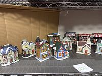 Front view of all 14 assorted ceramic Christmas village pieces arranged in a row showing various buildings including shops and houses with snow on roofs and holiday decor