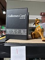 Closed black binder with silver text 'Collectors Card Album' and small Scooby Doo toy figure next to it.
