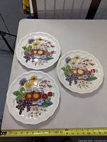 Three Copeland Spode Reynolds dinner plates with scalloped edges and fruit and flower motifs with grapes, pears, and various flowers arranged on a table with measuring tape showing about 9.5 inches in diameter.