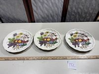 Three dinner plates with colorful fruit and floral designs and scalloped edges lined up on a flat surface with a ruler in front.