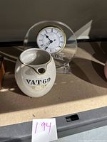 Cutty Sark desk clock with clear round acrylic base and white clock face with Roman numerals, next to Vat 69 Gold ceramic water pitcher on shelf