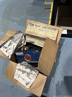 Open cardboard box containing various metal parts, including a blue perforated sheet, wheels, screws, and original instruction sheets for assembly.