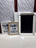 Three unopened items including one framed chalkboard with white frame and two unopened wood picture frames leaning against a backdrop.