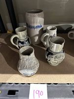 Four small German stoneware wine jugs with relief decorations and a tall stoneware beer stein with 'Maisingerbräu' label, displayed together.