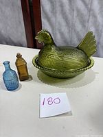 Green Indiana Glass hen covered candy dish next to two small colored glass bottles on white surface with handwritten lot number 180