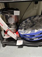 Lantern and soft cooler shown side by side on a black shelf with the lantern in an open box and cooler collapsed and wrapped in plastic.