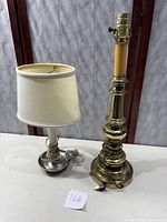 Photo showing two metal accent lamps on a white surface: a taller brass lamp without shade and a shorter silver-toned lamp with a white shade.