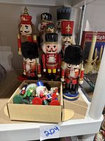 Eight larger nutcracker figurines arranged on a shelf with a box holding 10 smaller nutcracker ornaments in front, showing varied traditional soldier styles and painted details.