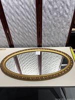 View of the full oval mirror on a white surface showing gold plastic frame and reflective glass.