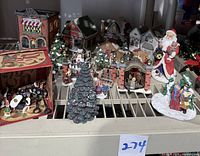 Photo showing porcelain buildings, small figurines, Christmas trees, and decorative accessories arranged on a shelf. Includes Gift Shop, Bentley Book Shop (chimney broken), Carriage House, Gift Store, and small resin Club House. Porcelain and resin figures visible.
