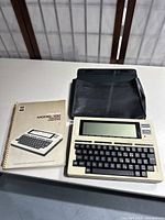 Full view of the TRS-80 Model 100 portable computer laid flat on a table alongside its user manual and black carrying case.