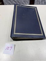 Blue binder with gold lined border closed on a table, label with number 127 nearby