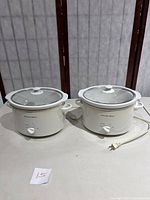 Two Proctor Silex white crock pots with glass lids on a table, showing front and side views.