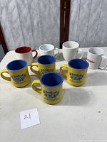 Group of eight coffee mugs showing four yellow Rainforest Cafe mugs with blue interiors, front row, and four assorted mugs behind