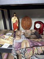 Photo showing Jesus statue, St. Thomas More Church commemorative clock, wooden carved cross plaque, angel throw blanket, necklace medallions, and small trinket dish on shelf