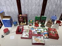 Full view of all Christmas holiday items including oil lamp, cards, votive holder, mug, keepsake box, and decorative sets on a white table with a folding screen behind it