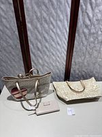 Full view of two Michael Kors cream tote bags and a blush wallet on a white table with a dark background.