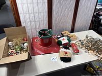 Full lot including metal tree stand, box of glass ornaments, green wreath, string lights, fabric door hangers, brass candlestick holders, and figurines on table.