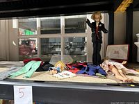 Overview of vintage Barbie and Ken dolls with multiple clothing pieces and Barbie jewelry chest under shelf