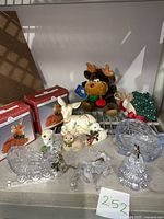 Overview of lot contents with porcelain deer, plush reindeer, glass reindeer and sleigh, Christmas ornaments, and other holiday decorations on a shelf.