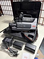 Full view of the vintage RCA VHS camcorder inside the open hard case, with two battery packs and charger placed in front.